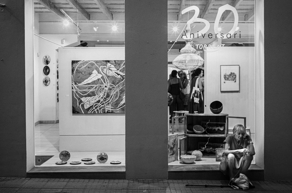 Sèrie en blanc i negre de la inauguració d’una exposició de Kike Mestre, amb visitants, el pintor, un ca i les obres dins la sala.
